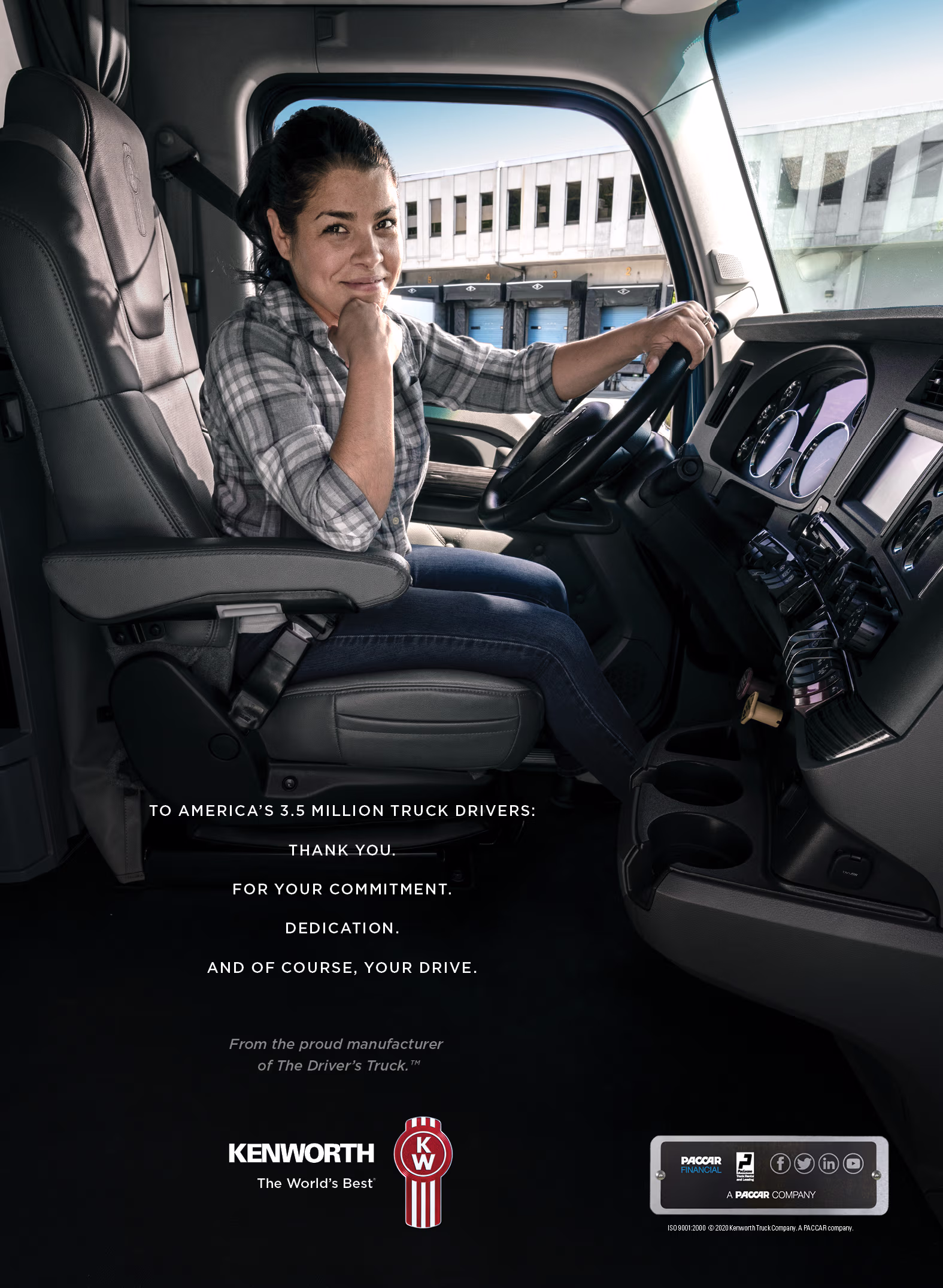 Truck driver in a plaid button-up shirt sits sideways in the driver’s seat of a Kenworth cab, resting her chin on her hand and looking toward the camera while holding the steering wheel; the cab interior and instrument cluster are visible, and a loading dock/building appears outside the window.